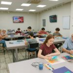 Group of people of various ages painting watercolors at long tables in a bright art classroom with supplies nearbyly visible in the background.