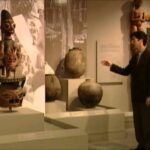 Man in a suit gestures toward ancient ceramic pots on display in a museum gallery.