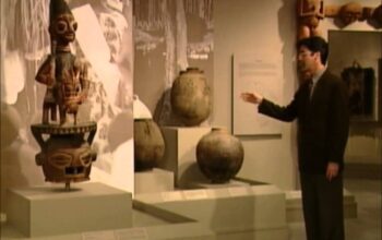 Man in a suit gestures toward ancient ceramic pots on display in a museum gallery.