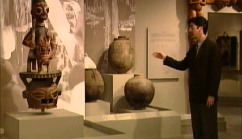 Man in a suit gestures toward ancient ceramic pots on display in a museum gallery.