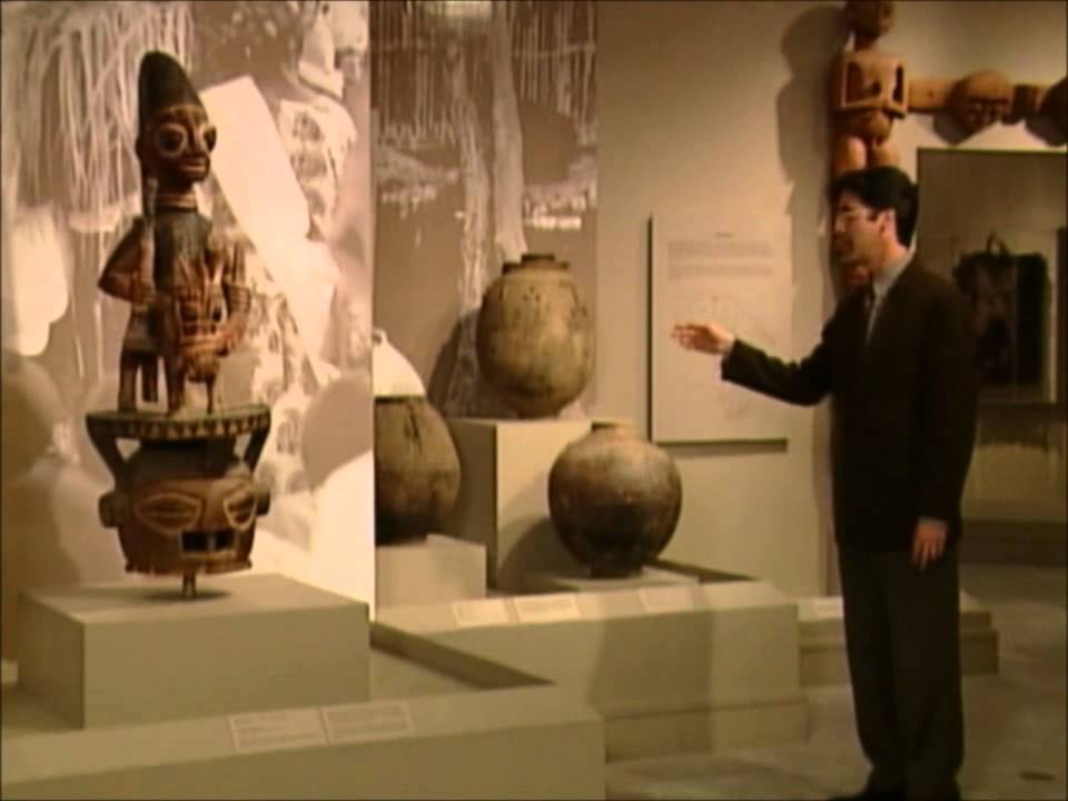 Man in a suit gestures toward ancient ceramic pots on display in a museum gallery.
